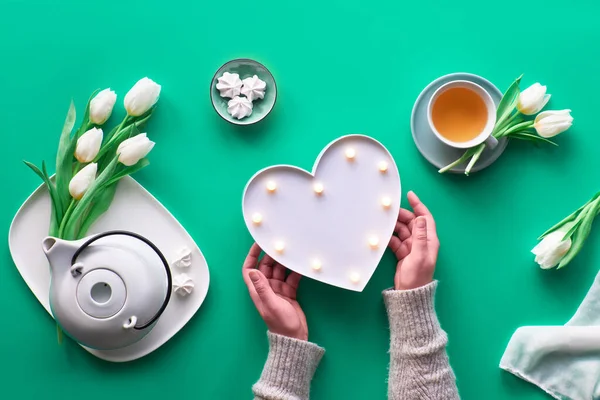 Frühlingsfest flach lag. Frauenhände zeigen Zeichen der Herzform. Teetasse, Teekanne, Süßigkeiten und weiße Tulpen auf grünem Tisch. Muttertag, Internationaler Frauentag, 8. März oder Mutti-Geburtstag. — Stockfoto