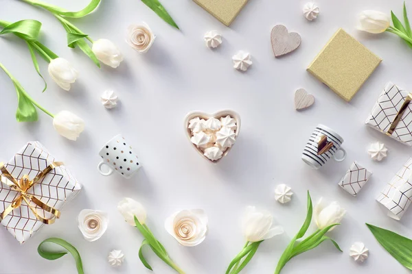 Valentijnsdag platte lay, bovenaanzicht op witte achtergrond met verse tulpenbloemen, koffiebekers, Valentijnscadeau in geometrische papier met gouden lijnen en marshmallows. Hartschaal met snoepjes in het midden. — Stockfoto