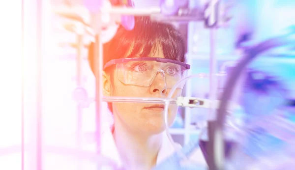 Young female scientist or tech in protective wear supervises chemical synthesis, checking reaction. Laboratory, glassware, futuristic orange and purple neon light, laboratory out of focus.