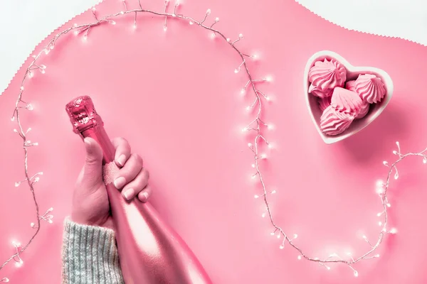 Vista dall'alto San Valentino su sfondo rosa. Ghirlanda leggera, mani di donna che mostrano il segno del cuore. bottiglia di champagne rosa metallico. Trendy monocromatico piatto giaceva in rosa vibrante con forme organiche astratte . — Foto Stock