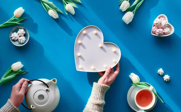 Happy Valentine or Mothers day celebration flat lay. Female hands show heart shape sign. Tea cup, tea pot, sweets and white tulips on tale, background in trendy classic blue color. — Stok fotoğraf