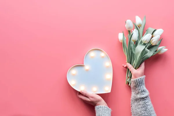 Springtime flat lay on pink, female woman hands holding bunch of white tulips, Spring flowers. Lightboard in heart shape in hands. Flat lay, top view, Mothers day or Valentine. — Stok fotoğraf