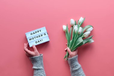 Bahar geometrik düzlük, ışık paneli ve pembe arka planda beyaz lale çiçekleri olan kadın elleri. Işık panosuna Mutlu Anneler Günü 'nü yaz. Kare kompozisyon. Anneler Günü kutlamaları.