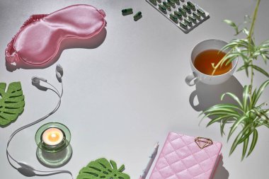Sağlıklı gece uykusu yaratıcı konsepti pembe ve yeşil. Uyku maskesi, kulaklıklar, çay, uyku hapları, günlük defteri. Dekoratif canavar yaprakları olan gri bir arka plan. Metin uzayı, kopyalama uzayı.