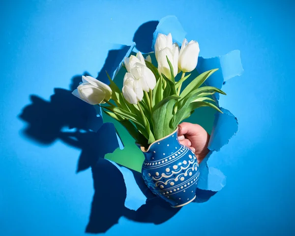 Happy Spring Holidays! Bunch of white tulips in classic blue color ceramic jug in hand shown through torn paper hole with long shadows. Trendy Spring Birthday, Easter, Mother\'s day holiday background.