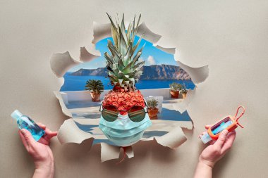 Covid-19 coronavirus sudden quarantine in remote tourist resort or hotel. Pineapple with sunglasses and medical face mask terrase and sea view through paper hole. Hands with disinfectant gel. clipart