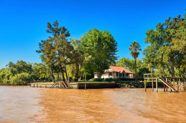 Tigra delta in Argentina, river system of the Parana Delta North from capital Buenos Aires. Lush vegetation, palm trees, wooden house under red roof. Orange clay water of Lujan River delta system. clipart