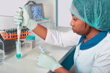 Koronavirüs için PCR testi. Beyaz önlüklü ve şapkalı Afrikalı genç bayan teknisyen, bilim adamı veya doktor hasta örneklerindeki SARS-CoV-2 'yi tespit etmek için testler yapar. Eldivenli ellerde otomatik pipet.