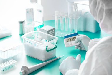 SARS-COV-2 PCR tanı kiti. Koruyucu giysi, maske ve gözlük takan epidemiyoloji uzmanı zamanlayıcı tutuyor, SARS-nCoV-2 virüsü tespit ediyor, Covid-19 viral zatürreye neden oluyor..