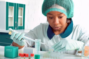 Koronavirüs için PCR testi. Beyaz önlüklü ve şapkalı Afrikalı genç bayan teknisyen, bilim adamı veya doktor hasta örneklerindeki SARS-CoV-2 'yi ve ağız örneklerini tespit etmek için test ediyor. Eldivenli ellerde otomatik pipet.