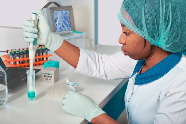 PCR test for coronavirus. African young female tech, scientist or medic in white coat and hat performs test to detect SARS-CoV-2 in patient samples. Automatic pipette in gloved hands.