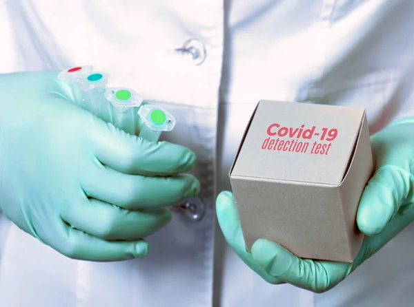 Medic or tech in white gown and gloves holds reagents for diagnostics of covid-2019 in patients with pneumonia. Test kit for pcr nucleic acid for presence of novel coronavirus, box and plastic tubes.