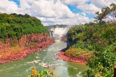 Arjantin 'de Iguazu şelaleleri. Turist motorlu teknesi güçlü su taşkınlarına doğru gidiyor ve Iguazu nehrinin üzerinde sis yaratıyor. Kırmızı taş kıyıları boyunca tropikal yağmur ormanlarının yemyeşil yaprakları...