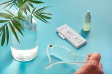 İgm ve IgG 'nin Corona virüsü SARS-CoV-2' yi geliştirerek Covid-19 hastalığına neden olan özel antikorların tespiti için hızlı COVID-19 testi. Mavi nane arkaplanı, palmiye bitkisi ve gözlük üzerine son sınıf el ve test.