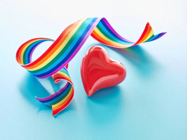 Red ceramic heart and rainbow ribbon, symbols of public support of medical teams fighting pandemics of novel coronavirus causing Covid-19 pneumonia. Curled rainbow ribbon on light mint blue background