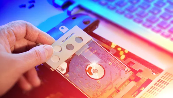 Hand with hard drive and electronics in hardware repair shop. Blurred image with opened laptop circuitry, close-up on electronics. Toned panoramic background, neon orange and blue light with flare.