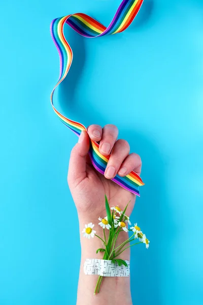 Thank you doctors and nurses! Rainbow ribbon in hand of mature woman with chamomile flowers and grass bouquet attached with medical patch. Creative flat lay, top view on blue background.