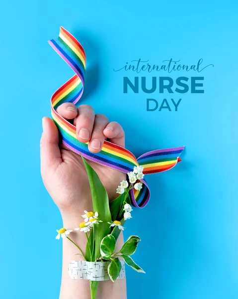 International nurse day concept. Hand of mature woman with chamomile flower bouquet attached with medical aid patch holding rainbow ribbon. Creative modern flat lay, top view on blue background.