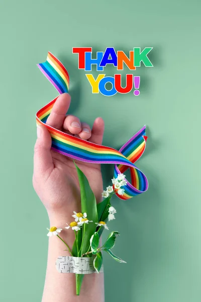 Rainbow ribbon in woman\'s hand. Chamomile flowers and grass bouquet attached to wrist with medical patch, aid band. Thank you doctors, nurses! Creative flat lay, top view on green background.