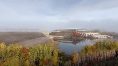 İnsansız hava aracı açık bir sonbahar gününde sakin bir nehirde kapalı bir baraja yaklaşır (Mactaqac barajı, New Brunswick, Kanada)