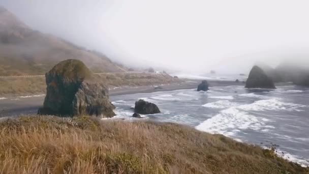 Cámara de drones se acerca a una costa montañosa en la niebla a través de acantilados en el agua Ariyas Beach, Oregon, EE.UU. — Vídeo de stock