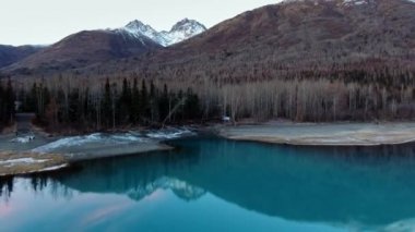 Drone, Alaska, Eklutna Gölü 'ndeki dağın eteğindeki ayna gölünün kıyısındaki kış ormanına yaklaşıyor.