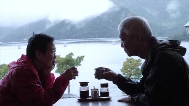 Азиатская старшая пара пьет чай вместе с красивым видом на горы и озеро в дождливый день — стоковое видео
