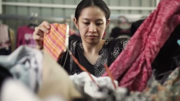Азиатская женщина смешанной расы покупает вторую одежду на складе блошиного рынка — стоковое видео