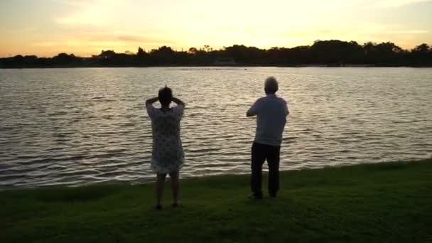 Asiático casal sênior exercitar juntos ao lado da luz da manhã com lago nascer do sol dourado — Vídeo de Stock