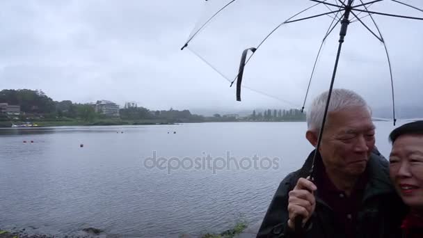 Casal asiático sênior sob guarda-chuva claro enquanto viaja para o lago nublado, explore o mundo juntos depois de se aposentar — Vídeo de Stock