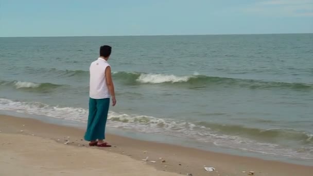 Азиатская старшая женщина, гуляющая одна вдоль пляжа, наслаждаясь природой — стоковое видео