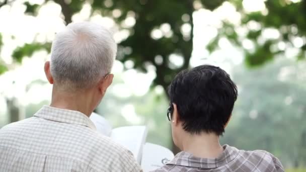 Vista trasera de la pareja de ancianos asiáticos sentados en el parque. Lectura de libro juntos en la atmósfera relax — Vídeos de Stock