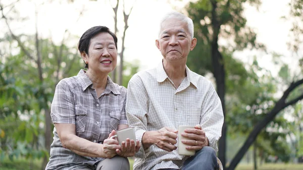 Old Asian senior couple morning date with cup of coffee green background park — Stock Photo, Image