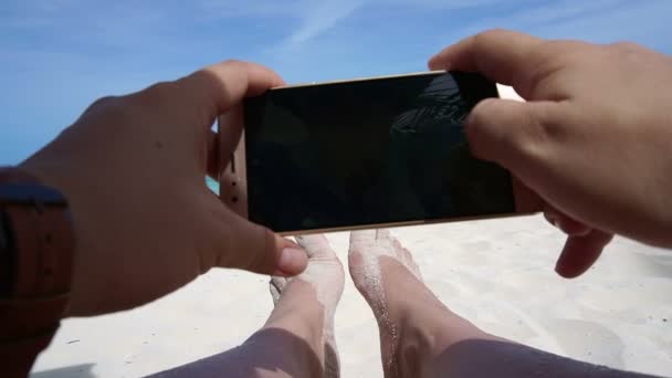 Άποψη του χέρι λήψη φωτογραφιών με έξυπνο τηλέφωνο. πόδια και ωκεανό φόντο διακοπές — Αρχείο Βίντεο