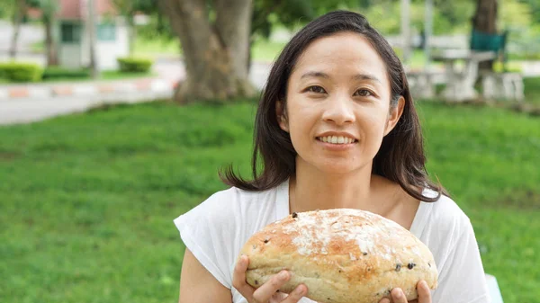 Asian woman holding and eating fresh baked bread bakery in green background park — Stock Photo, Image