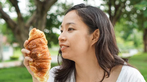 Asian woman holding and eating fresh baked bread bakery in green background park — Stock Photo, Image