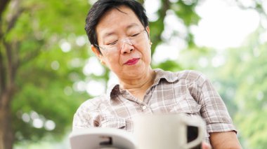 Asyalı kadın kıdemli güneşli park, kahve içme kitap okuma g