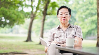 Asyalı kadın kıdemli güneşli park, kahve içme kitap okuma g