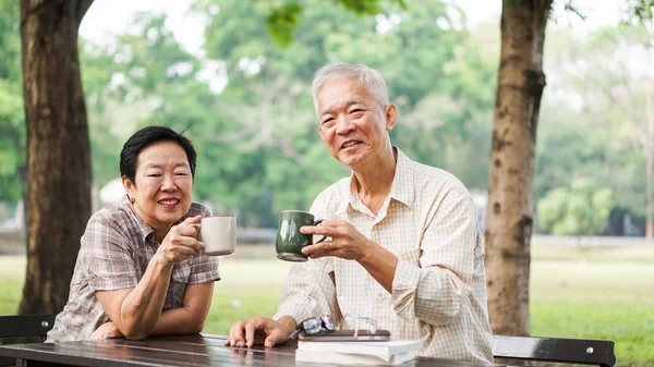 Asian senior couple relax drinking coffee in summer park, green — Stock Photo, Image