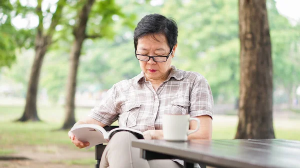 Asian senior woman reading book drinking coffee in sunny park, green background — Stock Photo, Image