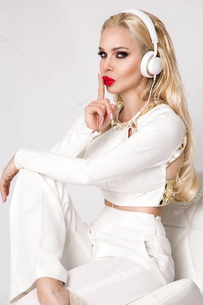 Beautiful sexy blonde woman with long hair and perfect body in an elegant white suit sitting with headphones on and listen to music 