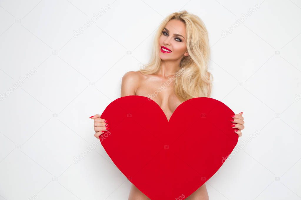 Beautiful woman with long blond hair standing on a white background dressed in red underwear and holds in his hands a red heart Valentine's Day