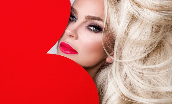 Vacker kvinna med långa blonda hår står på en vit bakgrund klädda i röda underkläder och håller i hans händer ett rött hjärta alla hjärtans dag — Stockfoto