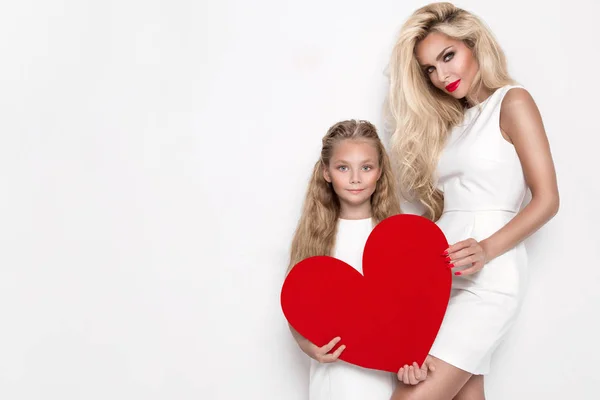 Όμορφη ξανθιά γυναίκα μητέρα και κόρη, στέκεται πάνω σε ένα λευκό φόντο και κρατώντας μια κόκκινη καρδιά στα χέρια του — Φωτογραφία Αρχείου
