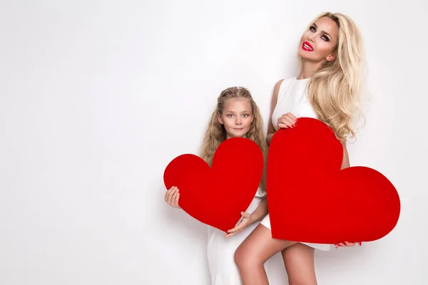 Hermosa mujer rubia madre e hija de pie sobre un fondo blanco y sosteniendo un corazón rojo en sus manos — Foto de Stock