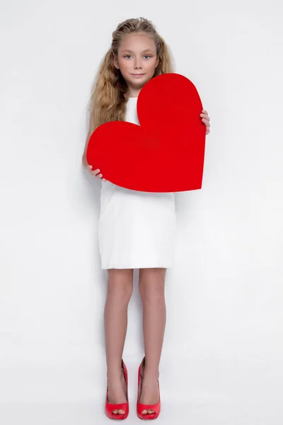 Bastante impresionante niña pequeña princesa, sosteniendo un corazón rojo en sus manos y de pie sobre un fondo blanco en el vestido blanco y zapatos rojos —  Fotos de Stock