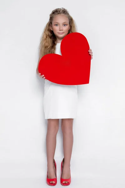 Pretty stunning little girl little princess, holding a red heart in his hands and standing on a white background in the white dress and red shoes — Stock Photo, Image