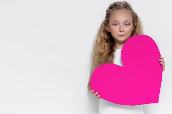 Docela ohromující holčička malá princezničko, hospodářství růžové srdce v ruce a stojící na bílém pozadí v bílých šatech — Stock fotografie