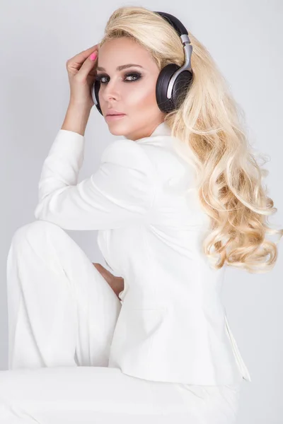 Mulher loira sexy bonita com cabelo longo e corpo perfeito em um terno branco elegante sentado com fones de ouvido e ouvir música e olhares sensuais — Fotografia de Stock