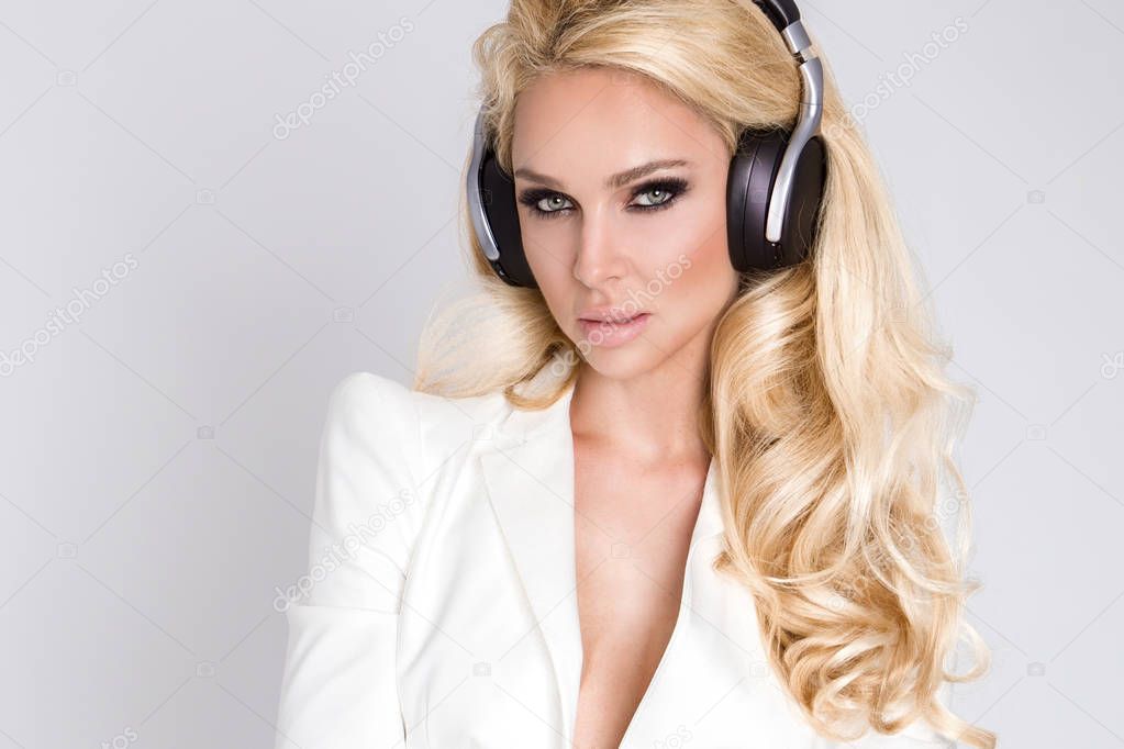 Beautiful sexy blonde woman with long hair and perfect body in an elegant white suit sitting with headphones on and listen to music and sensual looks
